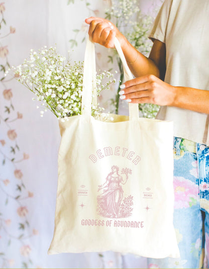 A cotton canvas totebag featuring various goddess designs including Demeter, Persephone, Aphrodite, Artemis, and Athena. The totebags come in shades of pink, gold, red, and black, of each goddess.